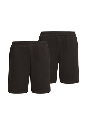 Personne portant un short noir mi-long, des chaussettes blanches montantes, des baskets blanches et grises, debout, mains dans les poches, sur un fond uni.