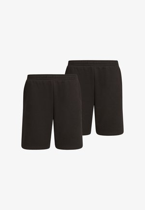 Personne portant un short noir mi-long, des chaussettes blanches montantes, des baskets blanches et grises, debout, mains dans les poches, sur un fond uni.