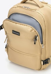 Mochila de nailon beige con cremalleras dobles, un bolsillo frontal y un interior gris. Cuenta con un parche de logo y correas ajustables. Textura suave.