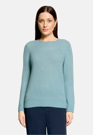 Femme aux cheveux bruns et raides portant un pull bleu clair à manches longues et un pantalon foncé, se tenant devant un fond clair uni.