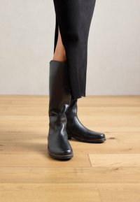 Gambe in una lunga gonna nera a coste con spacco laterale, che indossano stivali in pelle nera al ginocchio su un pavimento in legno.