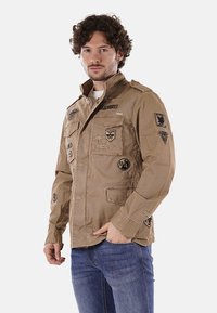 Giacca kaki con quattro tasche frontali, epaulettes sulle spalle e diversi patch ricamati. Polsini con bottone e chiusura frontale con zip.