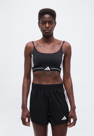 Sort kvinde iført sort Adidas sports-bh og shorts står mod en ensfarvet lys baggrund og vender fremad med et neutralt udtryk.