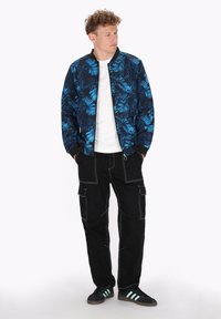 Bomberjack in marineblauw met een blauw palmenbladpatroon, gecombineerd met zwarte cargobroek met meerdere zakken en witte stiksels. Zwarte sneakers.