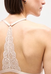 Γυναίκα με κοντά μαλλιά που φοράει ένα μπεζ σουτιέν racerback από δαντέλα, εμφανίζοντας λεπτομερή λουλουδάτο σχέδιο δαντέλας στο πάνω μέρος της πλάτης.