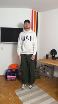 Witte hoodie met "GAP" logo, groene wijde broek en grijze sneakers. Staand op een gestreept vloerkleed, met een kleurrijke lamp en planten in de buurt.