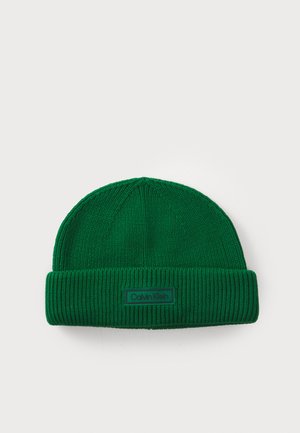 Green knit beanie featuring a ribbed cuff and a rectangular tag with the brand name. Textured fabric with a snug fit.