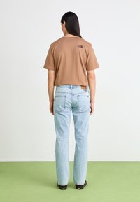 Korte bruine t-shirt met logo, gecombineerd met lichtblauwe rechtgesneden jeans. Achtergrond heeft een groene vloer. Schone en eenvoudige uitstraling.