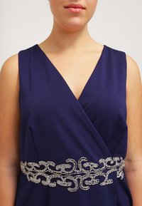 Námořnické modré šaty s V-výstřihem a kovově stříbrným korálkovým pásem s propracovanými vzory kolem pasu. Měkká textilie.