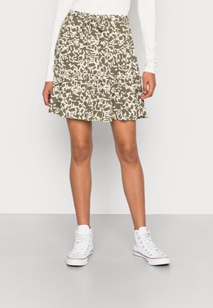 Ein gestufter, olivgrüner Rock mit einem weißen, abstrakten Muster, ausgestattet mit einem elastischen Bund und einem gerafften Saum. Gestylt mit weißen High-Top-Sneakern.
