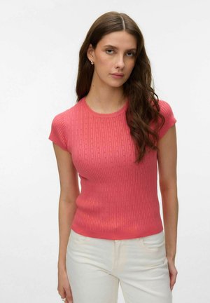 Jeune femme aux longs cheveux bruns portant un haut ajusté à manches courtes côtelé de couleur corail et un pantalon blanc cassé, devant un fond uni.