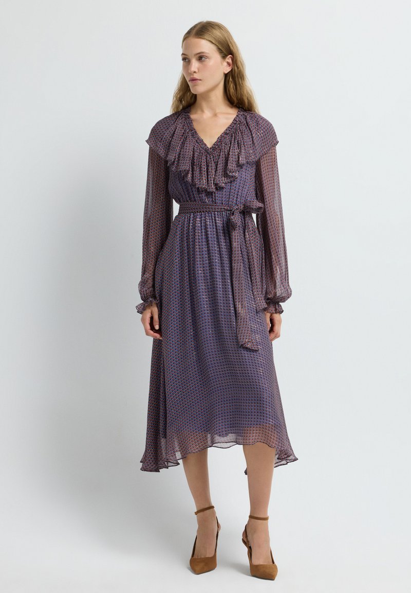 Vestido morado con estampado, escote en V con volantes, mangas largas y un cinturón en la cintura. La tela es transparente con una falda fluida. Zapatos de tacón alto marrones.