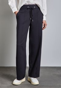 Pantaloni neri a gamba larga realizzati in un materiale morbido, con vita regolabile e tasche laterali, abbinati a sneaker bianche.