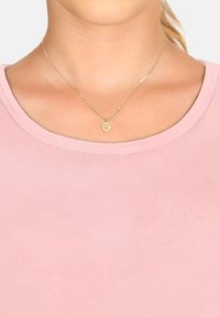 Collana d'oro con un pendente a forma di impronta di zampa, indossata su una morbida camicia rosa con scollatura rotonda. La collana presenta una catena delicata.
