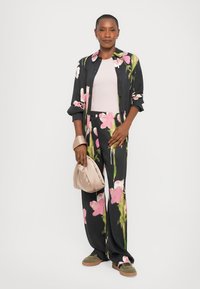Floral button-up shirt in black with pink and green accents, paired with matching pants. Model holds a light beige handbag and wears olive green shoes.