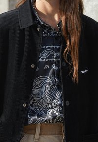 Sort fløjlsk jacket med trykknapper, båret over en marineblå satin skjorte med hvide paisley-mønstre. Lys beige bælte i udsigt nedenfor.