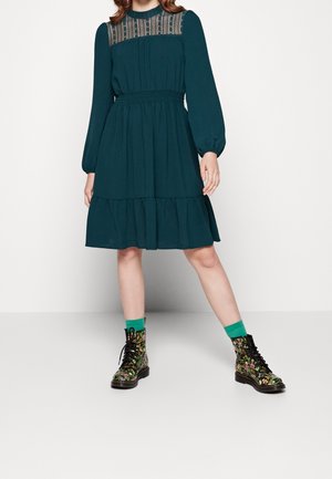 Turquoise lange mouw jurk met een kanten inzetstuk bij de hals, een gelaagde rok en een elastische taille, gecombineerd met combatlaarzen met bloemenpatroon en groene sokken.