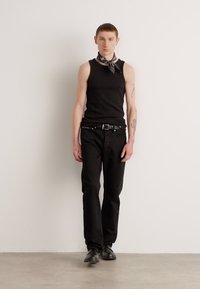 Črna brezrokavnik, črne jeans hlače in črne čevlje; dopolnjeno z vzorčasto ruto; model stoji ob enobarvni podlagi.