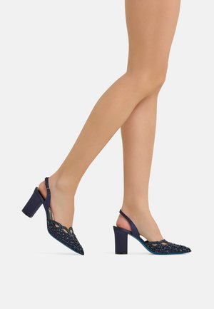 Beine mit marineblauen Slingback-Pumps mit Blockabsätzen und dekorativen Perlenverzierungen am spitzen Zehenbereich.