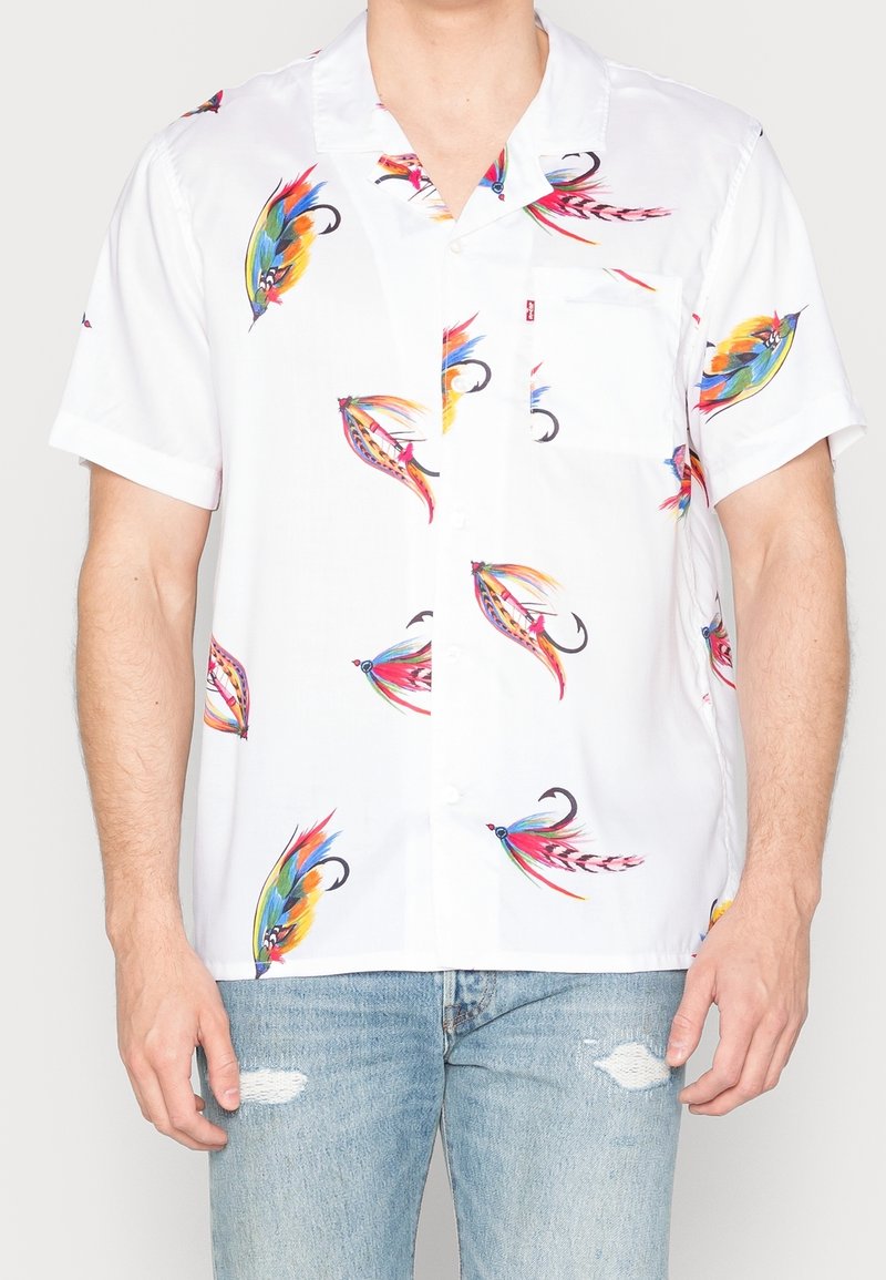 Biała koszula z krótkim rękawem z kolorowymi wzorami ryb, lekka tkanina, klasyczny kołnierzyk i kieszeń z przodu. Noszona z dżinsami typu distressed.