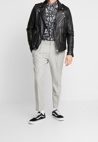 Giacca da motociclista nera in pelle, camicia a righe bianche e nere, pantaloni grigi chiari su misura, sneaker bianche e nere con lacci.