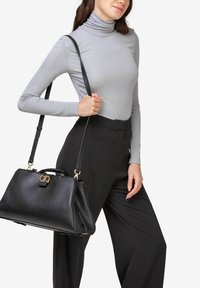 Sort læderhåndtaske med en struktureret form, gyldne hardware-detaljer og en aftagelig skulderrem, holdt af en person i en grå turtleneck.