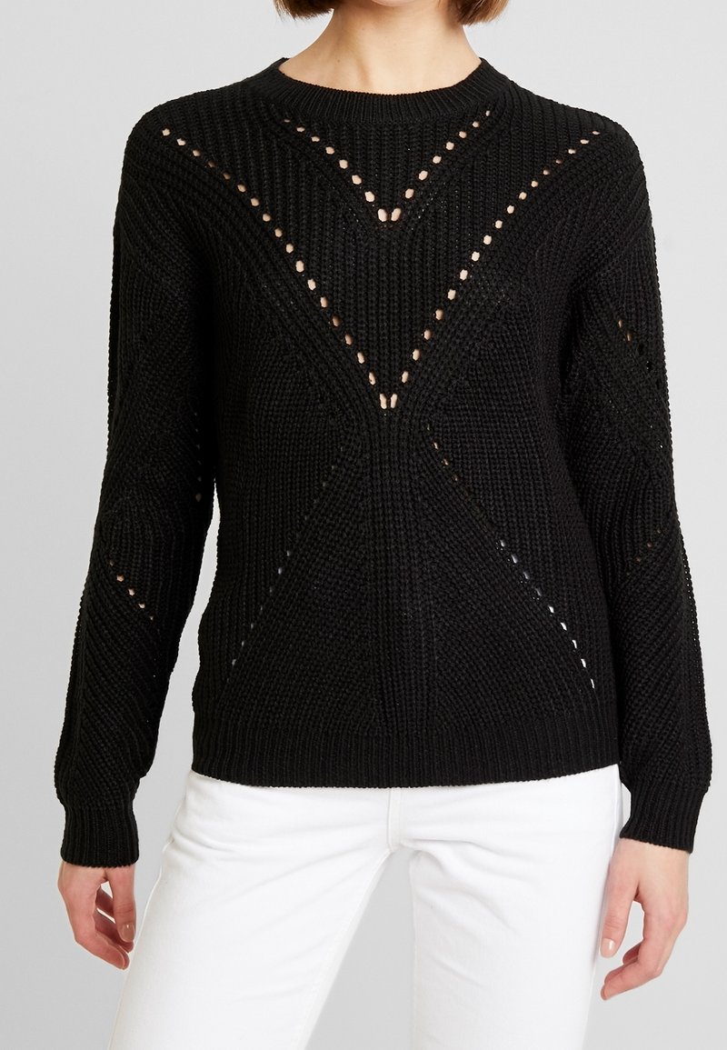 Schwarzer gestrickter Pullover mit einem strukturierten Muster und offenen Maschen, kombiniert mit weißen Jeans. Das Model steht vor einem grauen Hintergrund.