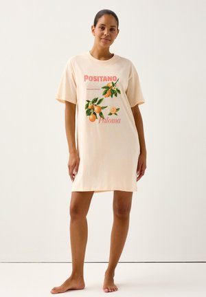 Moteris basomis stovinti, dėvinti šviesiai kreminės spalvos, per didelio dydžio marškinėlių suknelę su apelsinų vaisių ir lapų raštais bei užrašu „Positano Paloma“.