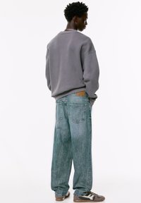 Sivi dukserica s spuštenim ramenima, uparena sa svijetloplavim, širokim trapericama koje imaju smeđi kožni detalj na struku. Smeđe tenisice.