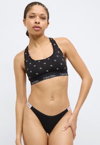 Μαύρο αθλητικό μπουστάκι Calvin Klein με μοτίβο λογότυπου και ελαστική ταινία, συνδυασμένο με μαύρο στρινγκ με λευκή λεπτομέρεια λογότυπου στη μέση.