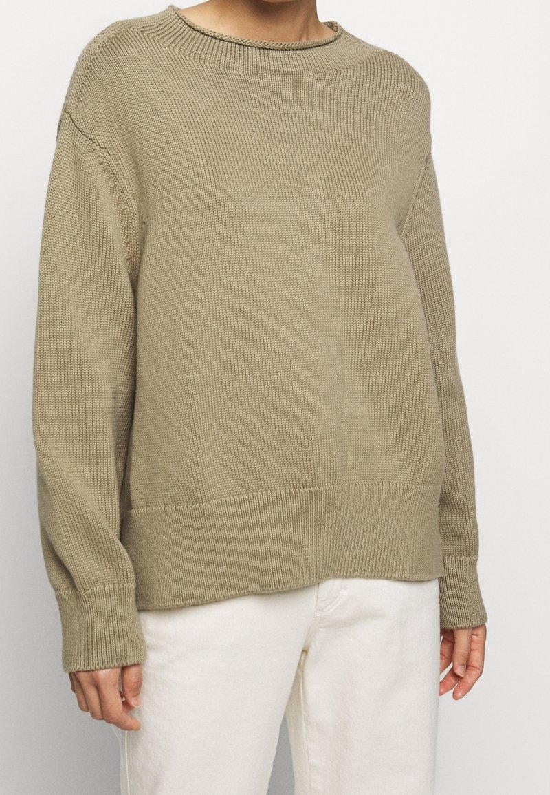 Personne portant un pull beige tricoté à manches longues avec des poignets et un ourlet côtelés, associé à un pantalon blanc cassé, sur un fond uni.
