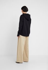 Felpa nera con cappuccio regolabile, abbinata a pantaloni beige a gamba larga, caratterizzati da una texture liscia e una forma rilassata.