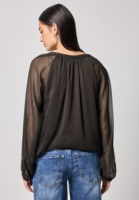 Schwarze, durchsichtige Bluse mit strukturiertem Muster, langen Ärmeln und gerafftem Rückendetail, kombiniert mit hellblauen Denim-Jeans.
