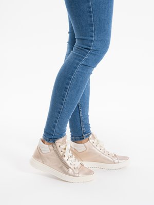 Legs wearing blue skinny jeans and metallic rose gold high-top sneakers with white laces and side zippers on a white background.