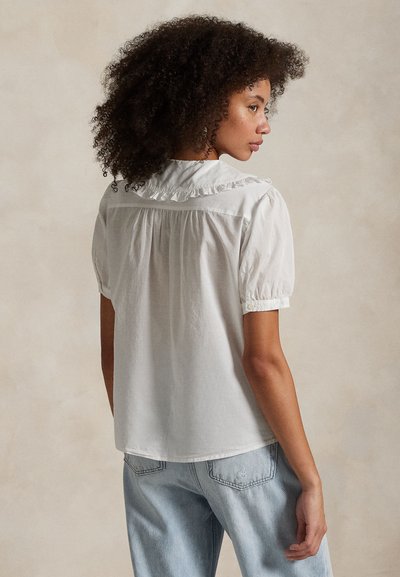 Kvinde med krøllet hår iført en hvid kortærmet bluse med flæsekrave og lyseblå jeans, set fra siden mod en ensfarvet baggrund.