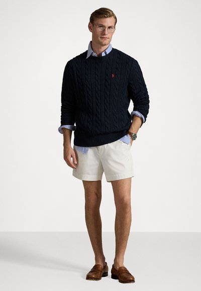 Marinblå kabelstickad tröja, ljusblå skjorta med krage under, beige shorts, bruna loafers. Enkelt mönster, texturerad tyg och klassiska färger.