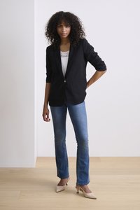 Žena s kudrnatými vlasy, která má na sobě černé sako přes bílý krajkový top, modré džíny a béžové boty s nízkým podpatkem, stojí na dřevěné podlaze.