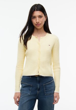 Vrouw met lang donker haar die een lichtgele, geknoopte cardigan en blauwe spijkerbroek met voorzakken draagt, staand tegen een witte achtergrond.