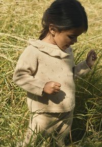 Pull en tricot beige avec un col à volants et de petits motifs, associé à un pantalon assorti, dans un champ herbeux.