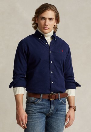 Jeune homme aux cheveux mi-longs portant une chemise bleu marine boutonnée sur un col roulé blanc, un jean bleu, une ceinture marron et une montre-bracelet, debout devant un fond neutre.