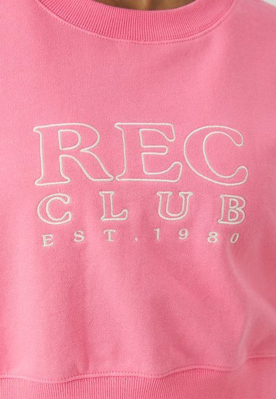 Rózsaszín pulóver bordázott nyakkal. Az elején fehér hímzett felirat található: "REC CLUB EST. 1980". Puha anyagú.