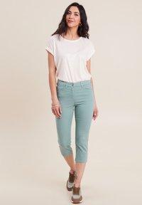 Top corto a maniche corte beige chiaro abbinato a pantaloni cropped verde menta. La modella indossa sneakers marroni e rosa. Design minimalista, vestibilità rilassata.
