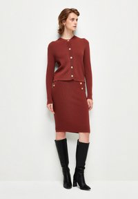 Cardigan a coste rossa con bottoni dorati, abbinato a una gonna al ginocchio dello stesso colore e stivali neri con tacco al ginocchio.