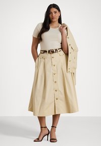 Gonna A-line beige, con bottoni, realizzata in cotone, con vita alta e tasche. Abbinata a un top fitted beige e a una giacca leggera.