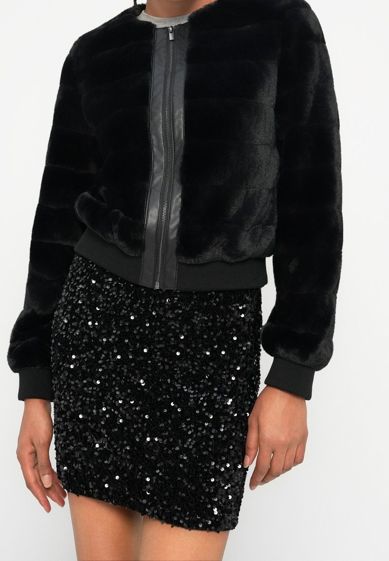 Personne portant une veste zippée en fausse fourrure noire et une mini-jupe noire à sequins sur un fond blanc uni.