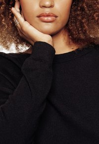 Primo piano del viso e del collo di un modello. Ha capelli castani ricci, labbra lisce con un finish lucido e indossa un maglione di lana nero.