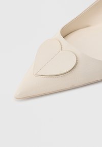 Sapato baile bege com bico pontiagudo e superfície texturizada, apresentando um destacado detalhe em forma de coração na parte frontal. Acabamento interior suave.