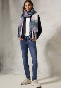 Unos jeans azules, una camiseta blanca y una bufanda multicolor con flecos. Las zapatillas presentan un diseño en beige claro y gris. Tejido suave, corte casual.