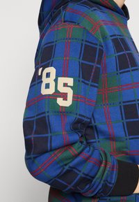 Sudadera a cuadros azul y verde con acentos rojos; presenta el detalle numérico '85' en la manga; textura suave y capucha ajustable.