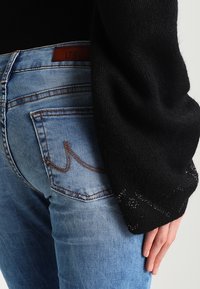 Jeans ajustados de mezclilla azul con un lavado desgastado, que presentan costuras de color cobre y una etiqueta de parche, combinados con un suéter negro holgado con mangas texturizadas.